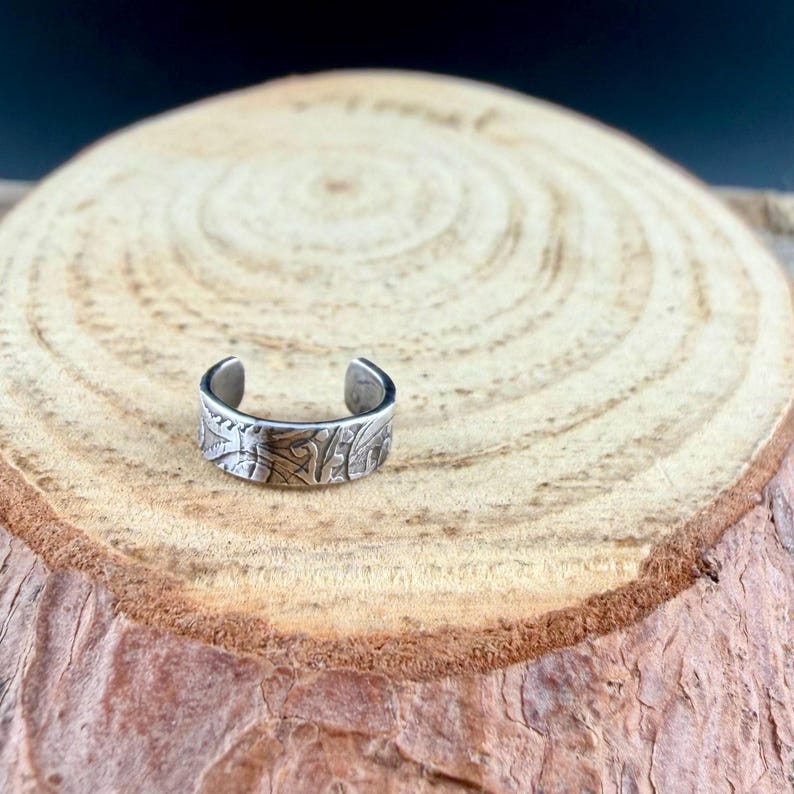 May include: A silver-colored, open-ended ring with an engraved floral pattern. The ring is displayed on a wooden surface with a natural, light brown grain. The ring's design features detailed, textured carvings.