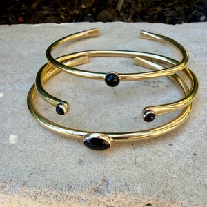 May include: Three gold-colored metal bracelets. Two are open-ended, and one has a black stone in the center. The third bracelet has two black stones on the ends and one in the center. The bracelets are stacked on top of each other.
