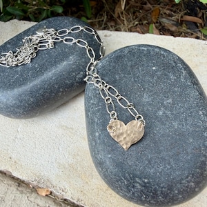 May include: A silver-toned necklace featuring a textured, hammered heart-shaped pendant suspended from a chain with elongated oval links. The metal surface of the heart displays a distinct dimpled pattern, reflecting light across its curved edges.