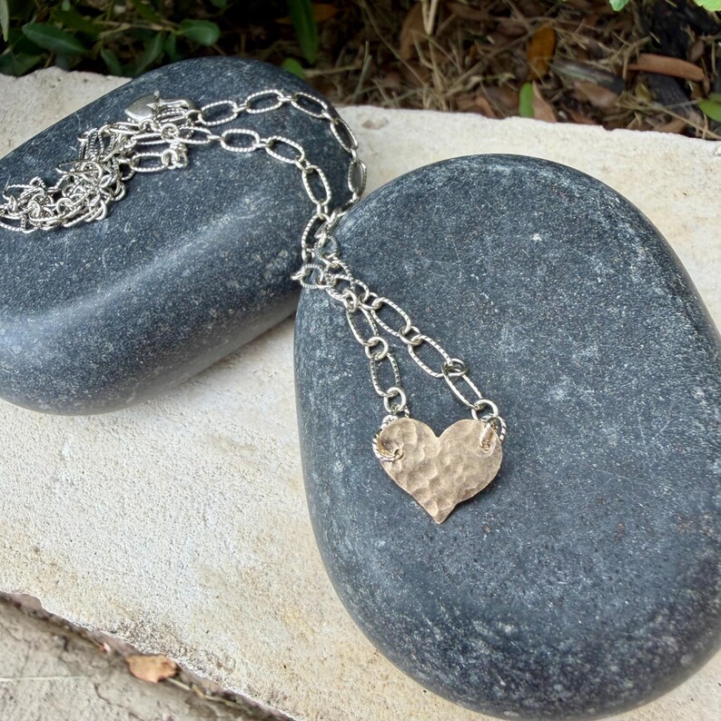 May include: A silver-toned necklace featuring a textured, hammered heart-shaped pendant suspended from a chain with elongated oval links. The metal surface of the heart displays a distinct dimpled pattern, reflecting light across its curved edges.