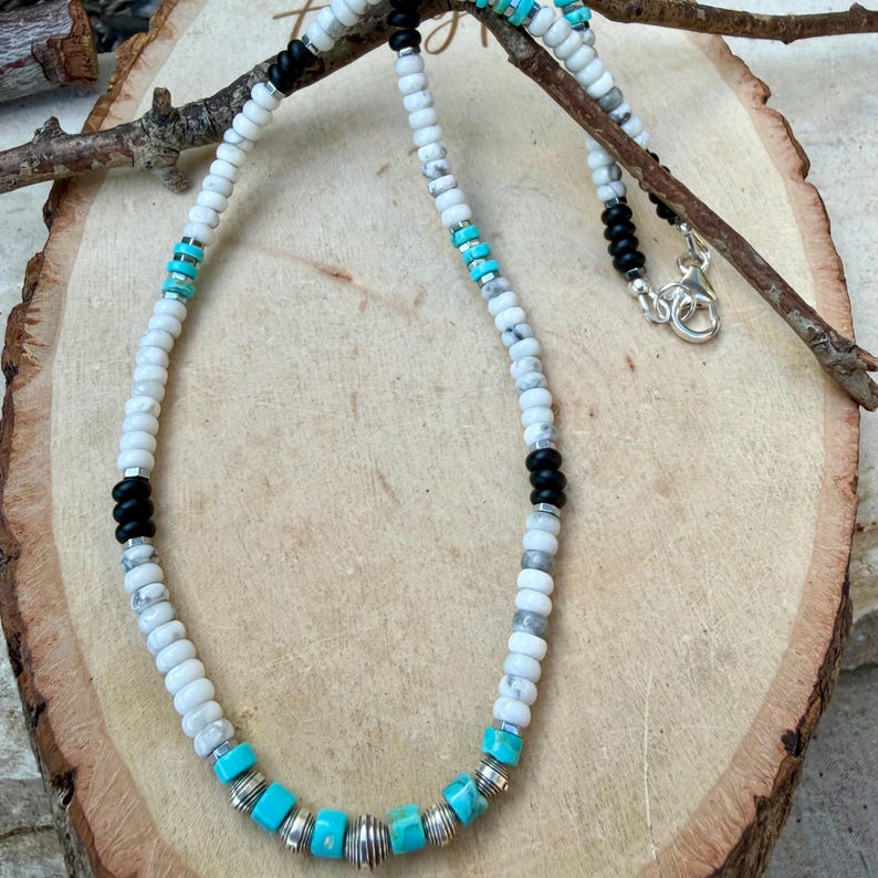 May include: A beaded necklace featuring white, black, and turquoise beads. The necklace has a silver clasp and is displayed on a wooden surface. The beads are arranged in a pattern, with turquoise beads interspersed with silver and white beads.