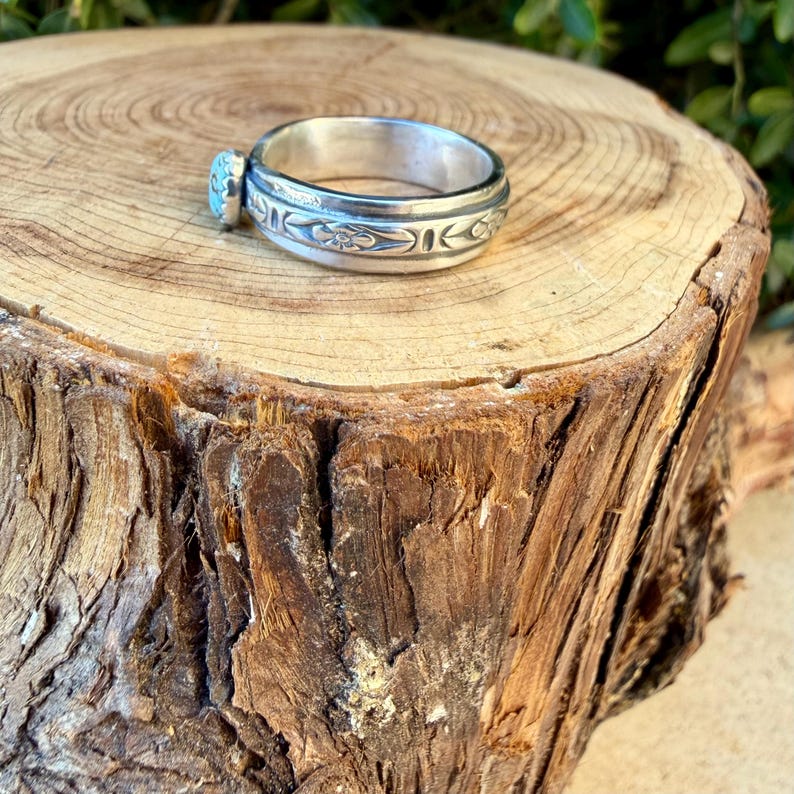 Puede incluir: Un anillo de plata con una banda pulida con patrones florales grabados y una peque&ntilde;a piedra redonda de color azul claro engastada en el lateral. El metal tiene un acabado brillante y reflectante, y la banda muestra un motivo decorativo detallado y uniforme.