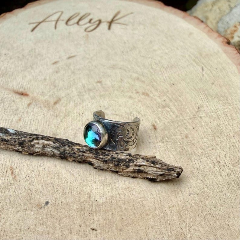 May include: A silver-toned ring with a round, iridescent blue-green stone. The ring band features an etched design. The word "Ally K" is etched into the wooden surface in the background. The ring rests on a piece of wood.