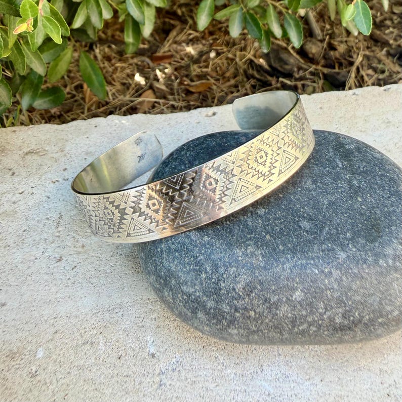 May include: A silver cuff bracelet featuring a repeating geometric pattern of etched triangles, diamonds, and lines. The metal band has an open back design and a polished finish, with visible markings on the inner surface near the opening.