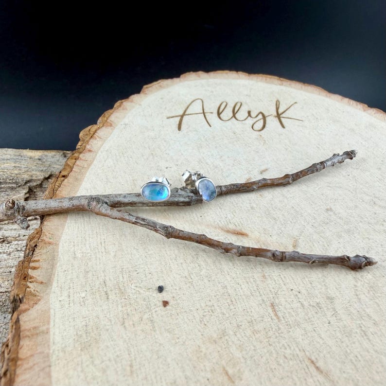May include: A pair of silver stud earrings with oval, iridescent blue gemstones. The earrings are displayed on a wooden slice with the handwritten text "Allyk" and a small twig.
