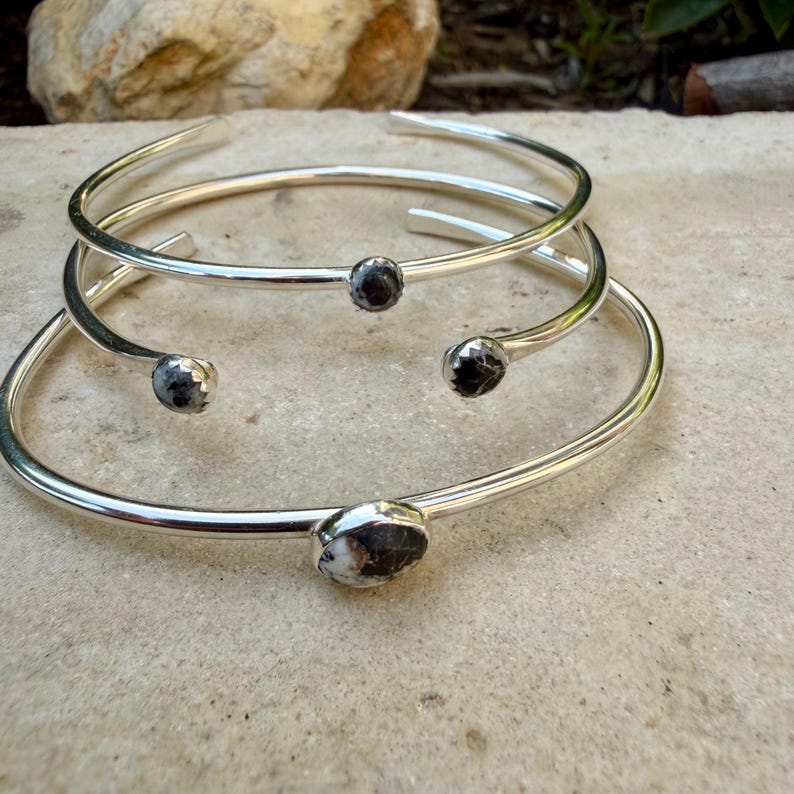 May include: Three silver-toned bracelets with dark, speckled stone accents. The bracelets are open-ended, with the stones set at the ends. The bracelets are displayed on a light-colored, textured surface.
