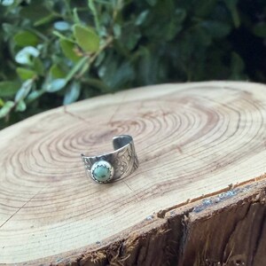 May include: A silver-toned cuff ring with an oval, pale green stone set in a textured bezel. The ring band features an etched pattern. The ring is displayed on a wooden surface with a natural wood grain.