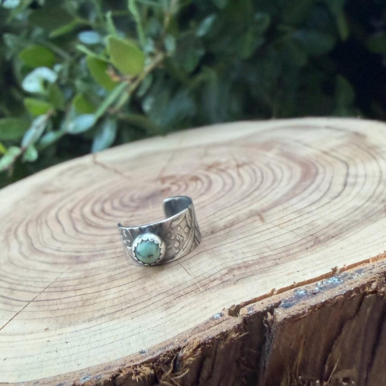 May include: A silver-toned cuff ring with an oval, pale green stone set in a textured bezel. The ring band features an etched pattern. The ring is displayed on a wooden surface with a natural wood grain.