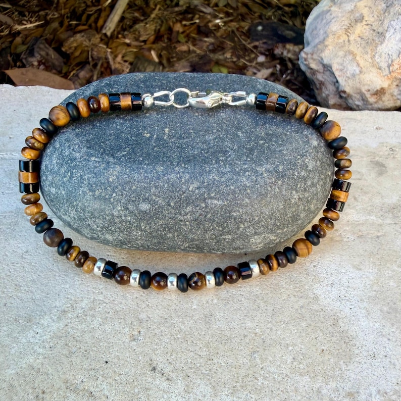 K&ouml;nnte beinhalten: Ein Perlenarmband mit abwechselnden runden Tigerauge-Perlen und kleinen schwarzen zylindrischen Perlen, akzentuiert durch silberfarbene Metall-Zwischenst&uuml;cke. Das Armband wird mit einem silberfarbenen Karabinerverschluss geschlossen.