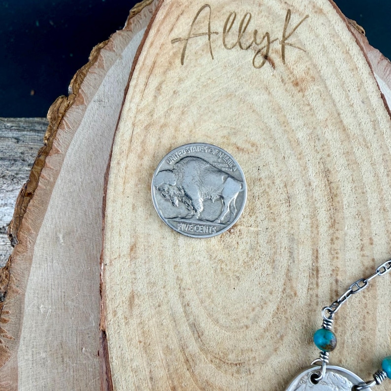 Op de afbeelding: Een zilveren Amerikaanse vijf cent munt met een buffelontwerp, geplaatst op een houten oppervlak. De munt ligt in de buurt van een ketting met turkooizen kralen. Het woord "Allyk" is in het hout ge&euml;tst.