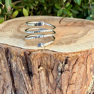 May include: A silver-colored metal ring featuring a spiral design with three distinct coils. The surface of the metal band has a hammered, textured finish, creating a reflective appearance across the continuous, open-ended coiled structure.