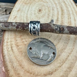 May include: A silver-colored ring with an intricate Celtic knot design, displayed on a wooden branch. A silver coin with a buffalo image and the words "FIVE CENTS" is also present, suggesting a rustic or handcrafted theme.