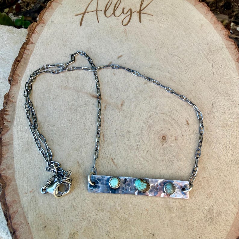 May include: A silver-toned necklace with a hammered metal bar pendant. The pendant features three turquoise-colored stones. The necklace is displayed on a wooden surface, with the handwritten text "Allyk" visible.