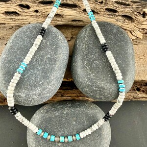 May include: A beaded necklace with white, black, silver, and turquoise beads. The necklace is displayed on gray stones and a piece of wood. The beads are small and round, with the turquoise beads being square-shaped.
