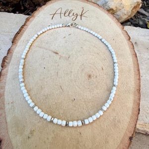 May include: White beaded necklace with silver accents and a clasp. The necklace is displayed on a wooden surface with the handwritten text "Allyk" in the upper left corner. The beads are small and evenly spaced.