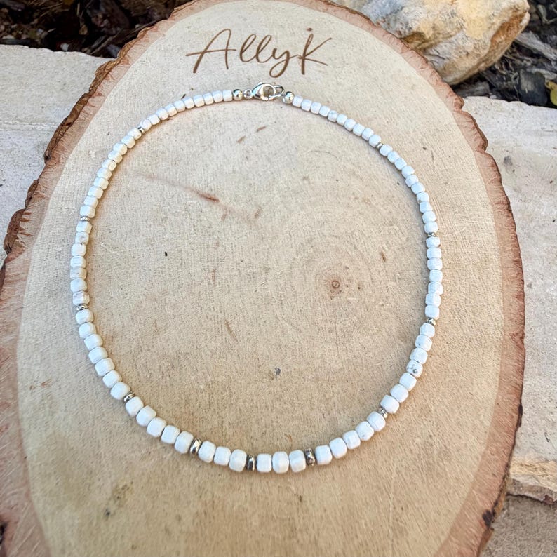 May include: White beaded necklace with silver accents and a clasp. The necklace is displayed on a wooden surface with the handwritten text "Allyk" in the upper left corner. The beads are small and evenly spaced.