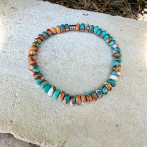 May include: A circular beaded bracelet featuring small, round, multi-colored stones in shades of turquoise, orange, white, and brown. The beads are arranged in a continuous loop, accented by a single, small, silver-toned metal spacer bead.