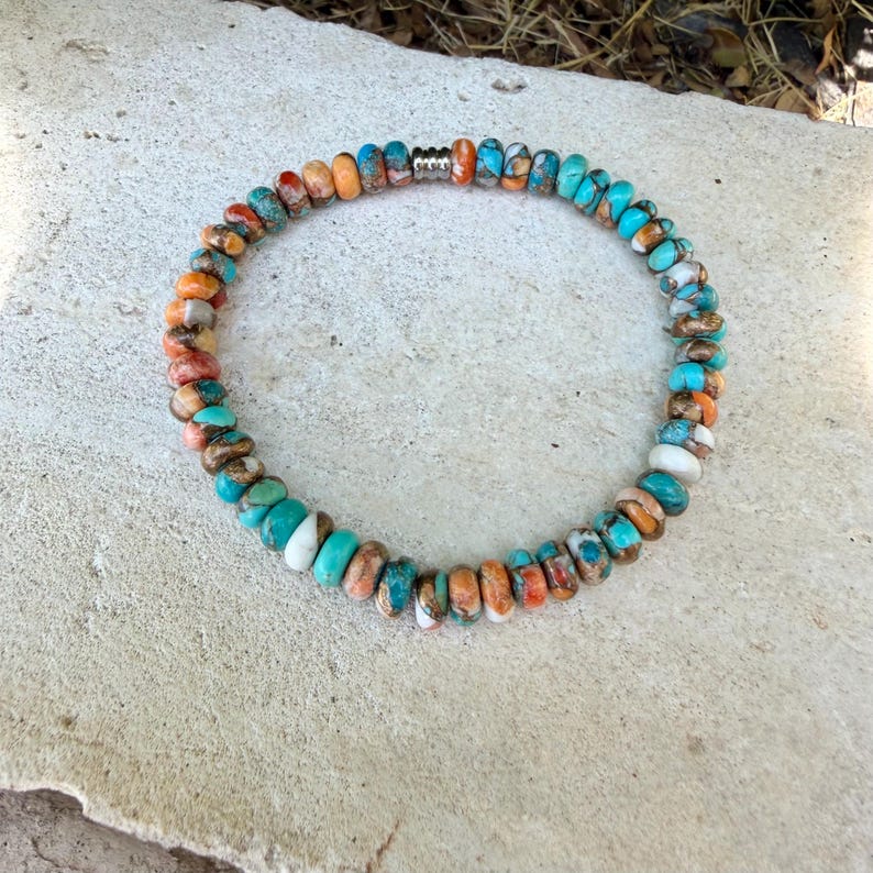 May include: A circular beaded bracelet featuring small, round, multi-colored stones in shades of turquoise, orange, white, and brown. The beads are arranged in a continuous loop, accented by a single, small, silver-toned metal spacer bead.