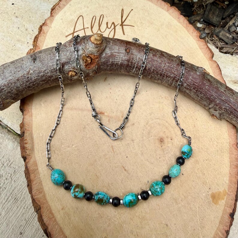 May include: A silver chain necklace with turquoise and black beads. The necklace is displayed on a wooden surface with the handwritten text "Allyk" in the upper left corner. The beads are a mix of round and oval shapes.