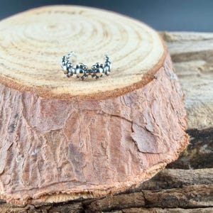 Puede incluir: Un anillo de plata para el dedo gordo del pie con un dise&ntilde;o floral. El anillo est&aacute; compuesto por peque&ntilde;os elementos redondos y texturizados dispuestos en forma curva. Descansa sobre una superficie de madera marr&oacute;n texturizada.