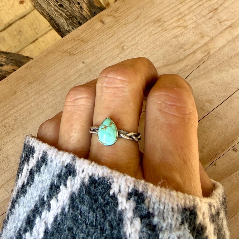 Puede incluir: Un anillo de plata con una piedra turquesa en forma de l&aacute;grima con vetas de matriz marr&oacute;n. La banda del anillo tiene una textura de cuerda trenzada y la piedra est&aacute; engastada en un bisel de plata liso. El anillo se lleva en un dedo.