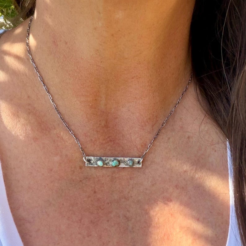 May include: A silver necklace featuring a rectangular pendant with three turquoise-colored stones. The pendant is attached to a delicate chain. The necklace is worn around the neck.