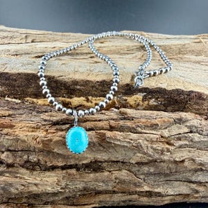 May include: A silver beaded necklace with a turquoise pendant. The pendant is oval-shaped and a bright turquoise color. The necklace is displayed on a piece of wood.