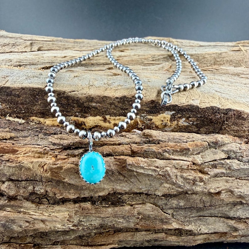 May include: A silver beaded necklace with a turquoise pendant. The pendant is oval-shaped and a bright turquoise color. The necklace is displayed on a piece of wood.