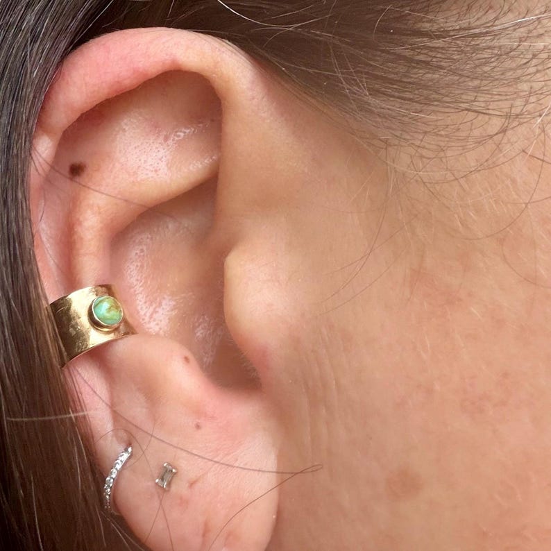 May include: A gold ear cuff with a round, light green stone is worn on the ear. A small, silver hoop earring with clear stones and a small, rectangular stud earring are also visible.