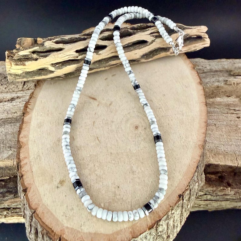 May include: White and gray beaded necklace with black accents. The necklace is made of small, round beads and features a silver clasp. The necklace is displayed on a wooden surface.