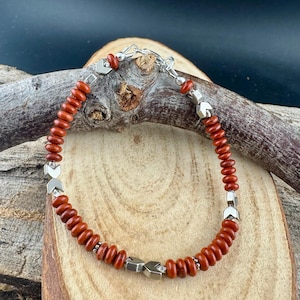 May include: A beaded bracelet featuring reddish-brown beads and silver-toned metal accents. The bracelet is strung on a silver chain and has a clasp closure. The beads are small and round, and the metal accents are geometric.