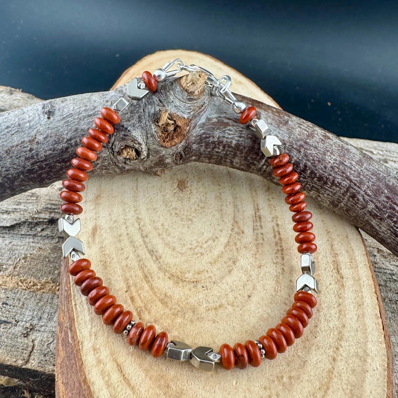 May include: A beaded bracelet featuring reddish-brown beads and silver-toned metal accents. The bracelet is strung on a silver chain and has a clasp closure. The beads are small and round, and the metal accents are geometric.