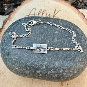 May include: A silver bracelet with a rectangular pendant featuring a cross design. The bracelet is made of a chain link design and is displayed on a dark gray stone. The word "Allyk" is written in cursive on a wooden surface.