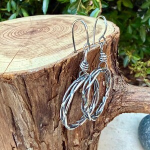 Op de afbeelding: Een paar zilverkleurige metalen oorbellen met een gedraaid, gevlochten draadontwerp in de vorm van een ovale ring. De oorbellen hangen aan eenvoudige gebogen oorhaken en hebben een gepolijste, reflecterende afwerking met textuur.