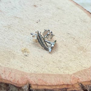 May include: A pair of silver feather stud earrings. The earrings are detailed with a feather design and have a post and butterfly backing. The earrings are displayed on a light brown wooden surface.