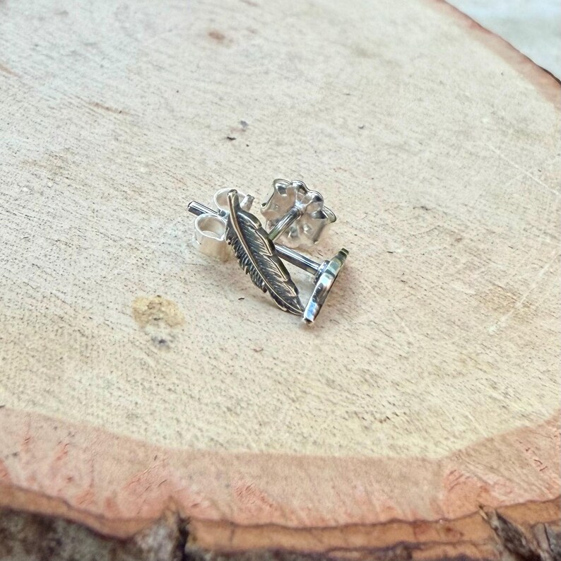 May include: A pair of silver feather stud earrings. The earrings are detailed with a feather design and have a post and butterfly backing. The earrings are displayed on a light brown wooden surface.