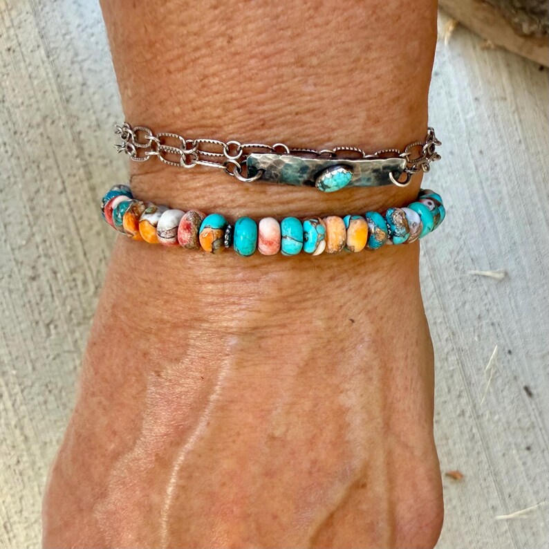 Pode incluir: Um conjunto de duas pulseiras composto por uma pulseira de corrente prateada com uma barra met&aacute;lica retangular central apresentando uma pequena pedra turquesa, e uma pulseira de contas redondas de pedra turquesa e laranja multicoloridas.