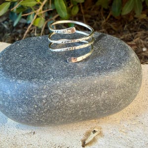 May include: A silver-colored metal ring featuring a multi-coil spiral design with a hammered, textured finish. The band consists of three overlapping loops that wrap around to form a continuous, open-ended spiral shape with rounded ends.