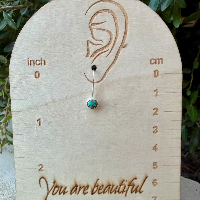 May include: A silver belly button ring featuring a round turquoise stone with brown veining. The jewelry is displayed on a light wood card with an engraved ear outline, inch and cm markings, and the text You are beautiful at the bottom.