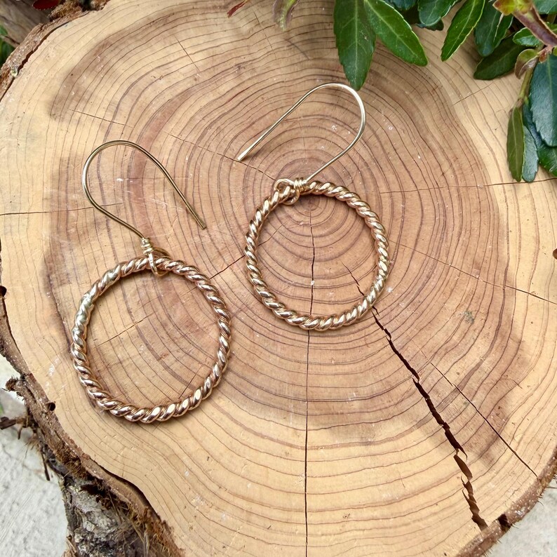 May include: A pair of gold-toned earrings featuring a circular hoop design with a twisted rope texture. Each earring is suspended from a curved ear wire hook, creating a simple and cohesive look for these metallic circular jewelry pieces.