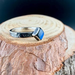 May include: A silver ring with a square, blue gemstone. The band features an etched leaf design. The ring is displayed on a wooden surface with a dark background.