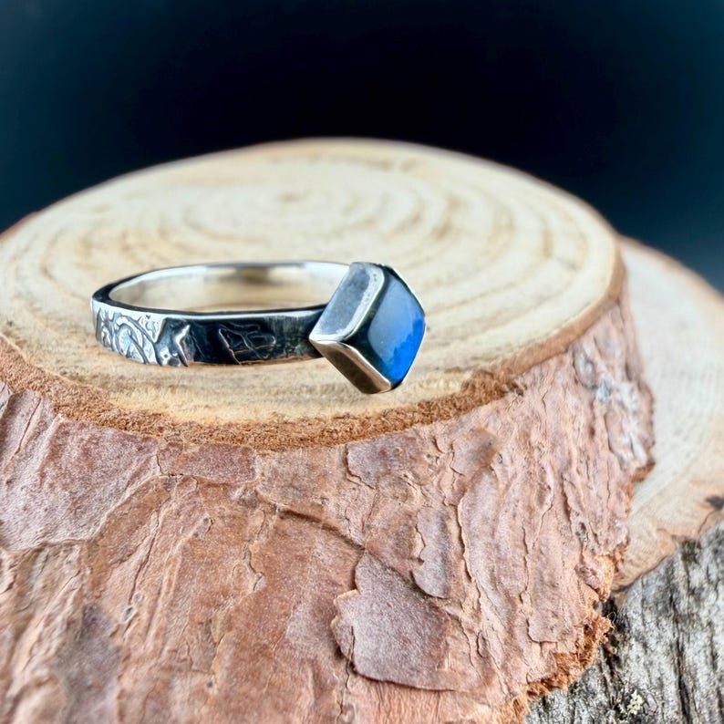 May include: A silver ring with a square, blue gemstone. The band features an etched leaf design. The ring is displayed on a wooden surface with a dark background.