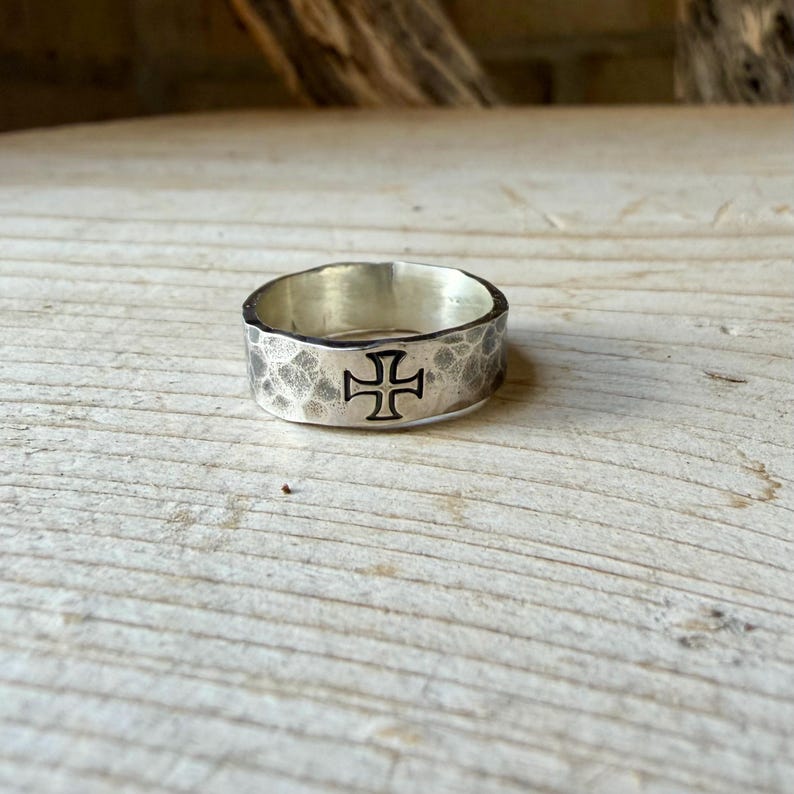 May include: A silver band ring featuring a hammered, textured finish and a prominent black engraved cross symbol centered on the outer surface. The metal has a polished interior and a rustic, uneven exterior texture throughout the entire band.
