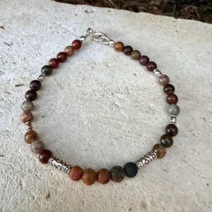 May include: A beaded bracelet featuring round, multi-colored earth-toned stones in shades of brown, gray, and tan. The bracelet is accented with small, cylindrical silver-toned metal spacers and a silver-toned lobster claw clasp closure.