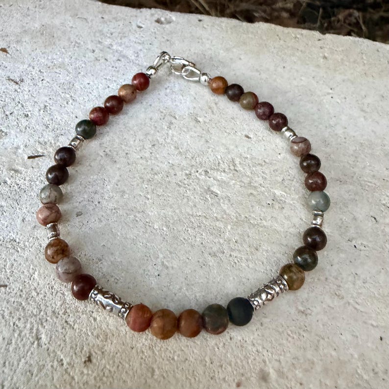 May include: A beaded bracelet featuring round, multi-colored earth-toned stones in shades of brown, gray, and tan. The bracelet is accented with small, cylindrical silver-toned metal spacers and a silver-toned lobster claw clasp closure.
