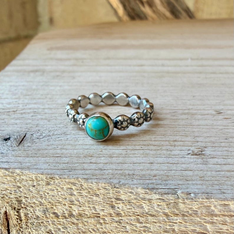 以下が含まれることがあります： トルコ石の付いたシルバーリング。リングは、小さな丸いディテールと中央のトルコ石が付いたテクスチャード加工されたバンドです。トルコ石は鮮やかで鮮やかな青緑色です。リングは木製の表面に置かれています。