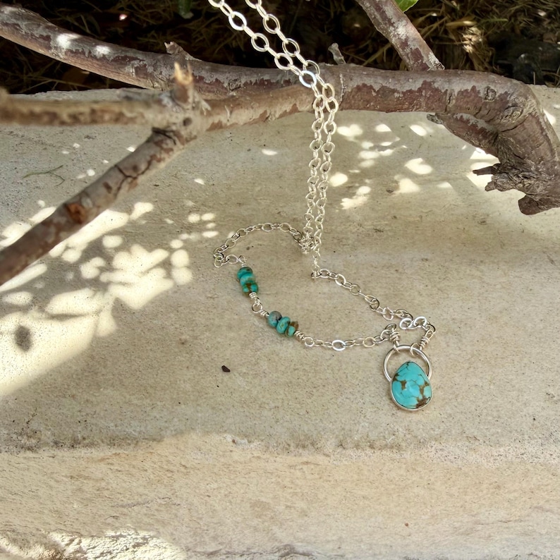 May include: A silver chain necklace featuring a teardrop-shaped turquoise pendant and small turquoise beads integrated into the chain links. The necklace has a metallic silver finish and displays natural blue-green stone variations throughout.