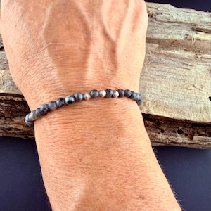 May include: A beaded bracelet featuring small, round, grey and black mottled stone beads interspersed with polished silver-toned metal spacer beads. The bracelet is strung on a flexible cord and fits snugly around the wrist.