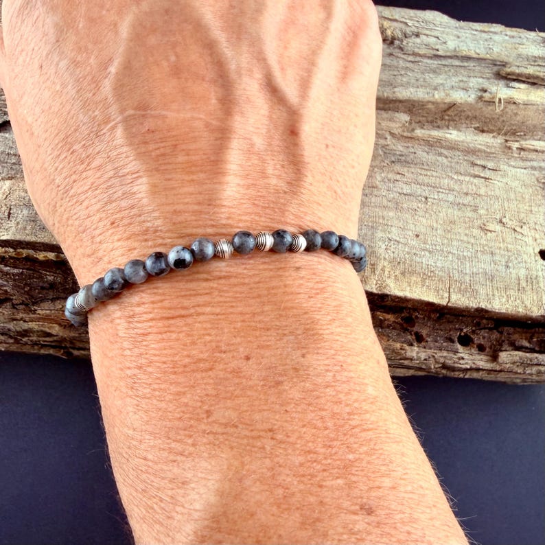 May include: A beaded bracelet featuring small, round, grey and black mottled stone beads interspersed with polished silver-toned metal spacer beads. The bracelet is strung on a flexible cord and fits snugly around the wrist.