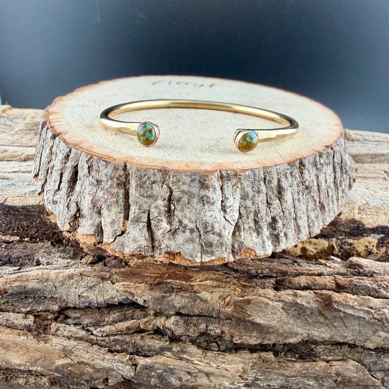 May include: A gold-colored cuff bracelet with turquoise-colored stones at each end. The bracelet is displayed on a rustic wooden slice, showcasing its design and texture. The bracelet is a piece of jewelry.